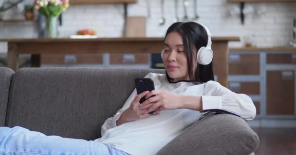 Sereno jovem mulher asiática relaxante no sofá confortável com os olhos fechados usando fones de ouvido. Bonita senhora gosta de ouvir música fria som de áudio meditando sentindo nenhum estresse em casa. Close up vista lateral — Vídeo de Stock