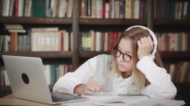 Kulaklık takan ve gözlük takan genç bir kız uzaktan öğretmeniyle internetten ders çalışıyor. İnternette öğrenim gören, notlar alan, ödev yapan, video dersi izleyen gençler.