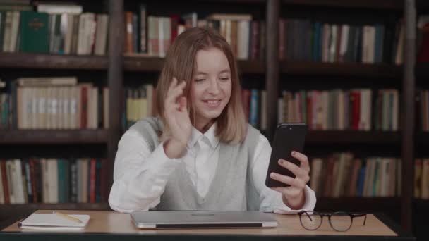 Smart tonåring flicka skolan elev viftar hand med hjälp av video konferens samtal via webbkamera på smartphone bibliotek i bakgrunden. Utbildning på videokonferens — Stockvideo
