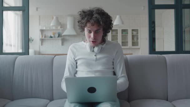 Een jonge krullenbol die thuis haar laptop gebruikt. Bank in witte kamer. Man op zoek scherm laptop en zeggen JA. win concept — Stockvideo