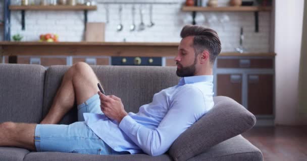 Молодий чоловік тримає пристрій смартфона смс-повідомлення, лежачи в дивані в домашньому офісі, усміхнений хлопець використовує програми, які грають в мобільні ігри балачки в соціальних мережах, серфінг в Інтернеті на телефоні в приміщенні — стокове відео