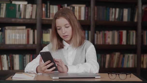 Criança loira jovem com cabelo curto bem vestido usando telefone celular na biblioteca da escola, conversando fofocas ou uma mensagem de voz com o amigo — Vídeo de Stock