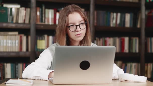 Positief kind in bril en met behulp van laptop thuis bibliotheek studeren online. Zijaanzicht van schoolmeisje in koptelefoon kijken online les op laptop en het opschrijven van notities. E-learning-concept — Stockvideo