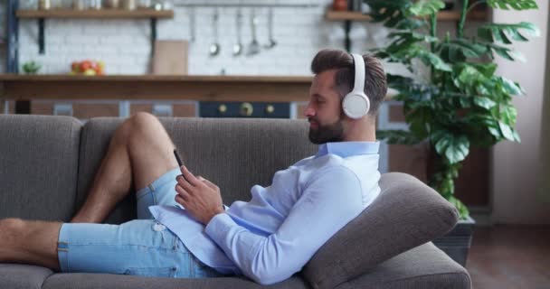 Glad hipster kille i att bära moderna trådlösa hörlurar, välja att njuta av favoritmusik från mobilapplikation, koppla av ensam på bekväm soffa i vardagsrummet. — Stockvideo