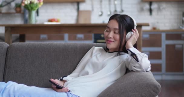 Mujer atractiva asiática escuchando música con auriculares que sostienen el teléfono inteligente encendido, sentado en el sofá en el apartamento cómodo — Vídeos de Stock