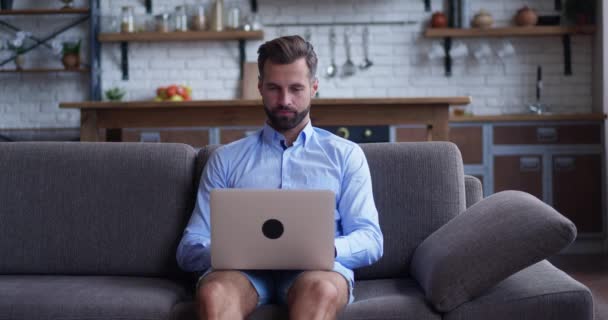 Γενειοφόρος νεαρός άνδρας κάθεται σε έναν καναπέ κρατά φορητό υπολογιστή στην αγκαλιά του, Περιηγήσεις μέσω του Διαδικτύου, Κοινωνικά Δίκτυα, Κάνει e-Shopping. Ο άνθρωπος στο σπίτι χρησιμοποιώντας Laptop, ενώ κάθεται σε έναν καναπέ. — Αρχείο Βίντεο