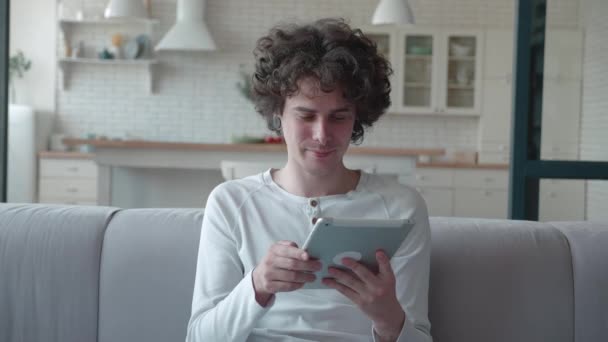 Молодой человек использует планшетный компьютер, сидя дома на диване. Хипстер с кудрявыми волосами ходит по Интернету и улыбается — стоковое видео