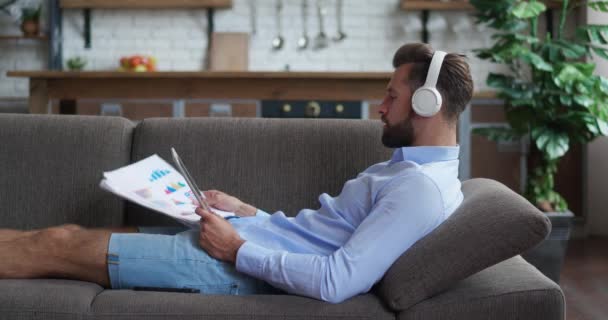 一个戴耳机的英俊商人躺在舒适的沙发上，正在做数字平板电脑的文档纸 — 图库视频影像