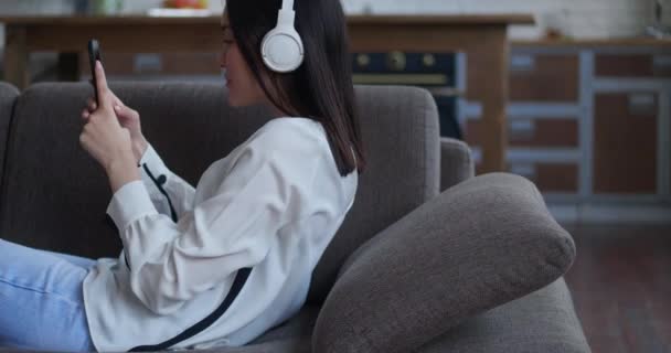 Feliz mulher asiática ouvir músicas usando fones de ouvido segurando smartphone tocando tela deitada no sofá aconchegante em casa. Enquanto relaxa no sofá em casa — Vídeo de Stock