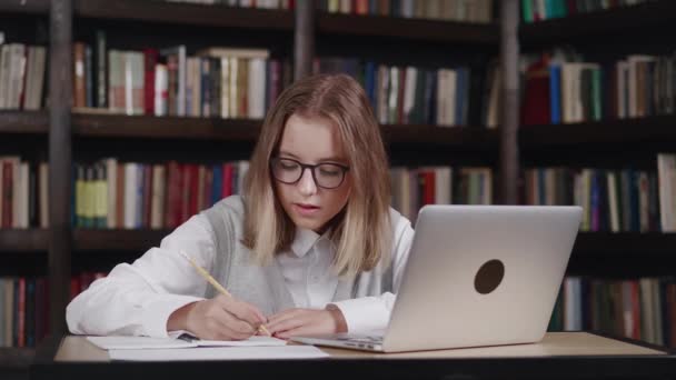 Menina vestindo óculos fazendo lição de casa estudante estudando on-line usando laptop fazendo anotações no caderno enquanto tem uma chamada de vídeo com professora no laptop na biblioteca — Vídeo de Stock