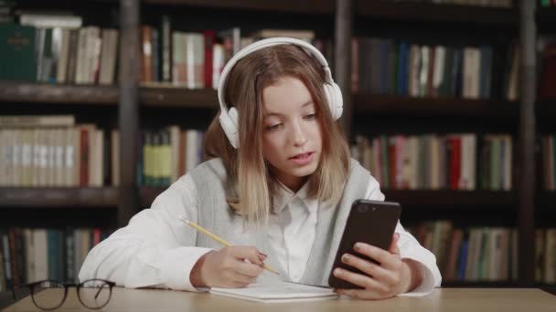 Het schoolmeisje met hoofdtelefoon geeft les terwijl ze thuis zit in de keuken aan tafel met behulp van een smartphone, boeken, notitieboekje. Close-up — Stockvideo