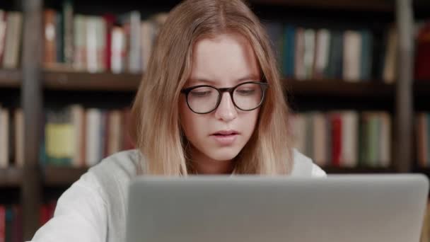 Sluit een jonge blonde tiener meisje in glazen werken op de computer op school bibliotheek. Casual geconcentreerde vrouwelijke het ontwikkelen van nieuw project tijdens het werken op laptop. Onderwijsconcept — Stockvideo