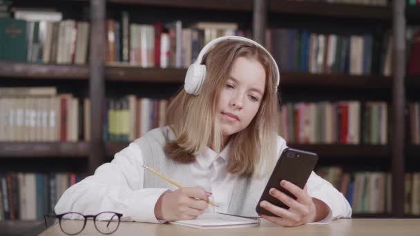 Roztomilé dítě dívka nosí sluchátka studující on-line v mobilní aplikaci videohovory se vzdáleným učitelem, vychovatel. Školák, který drží telefon, dělá domácí úkoly pomocí aplikace, dívá se na třídní učení doma. — Stock video
