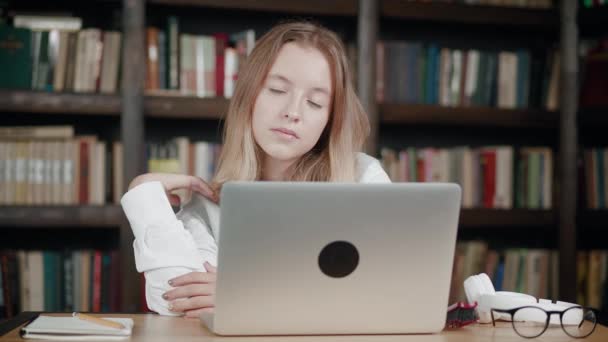 Portret. Uczennica robi pracę domową za pomocą laptopa, siedząc w bibliotece domowej i patrząc w aparat z uśmiechem — Wideo stockowe