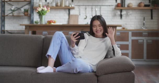 Menina asiática deitada no sofá ter vídeo chat no smartphone conversa casual em casa. Uma jovem atraente a ligar, a falar na câmara do telemóvel, a sorrir e a rir. Conceito de auto isolamento. — Vídeo de Stock