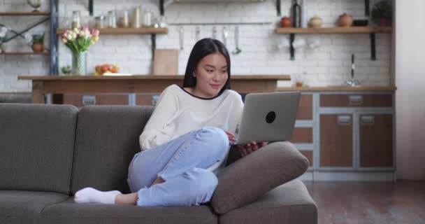 Freelancer asiático en camisa blanca acostado en el sofá con el ordenador portátil, tecleando en el teclado y parpadeando mientras mira la pantalla — Vídeos de Stock