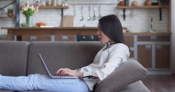 Joven mujer asiática freelancer acostado en el sofá en casa y trabajando en el ordenador portátil. Sentirse emocionado de escuchar noticias increíbles, celebrar el éxito o ganar el concepto de buena suerte. — Vídeo de stock