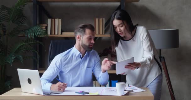 Un homme d'affaires et une assistante asiatique discutent de données financières analysant les résultats du marketing à l'aide d'une tablette numérique au bureau et collationnent un rapport à l'aide d'un ordinateur portable ayant une conversation d'affaires — Video