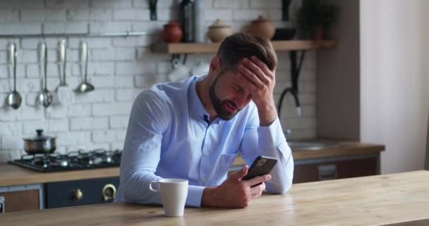 Frustrasi pada pria sedih yang tampak seperti smartphone. Karyawan yang tertekan membaca berita buruk di smartphone di dapur modern. Kesepakatan yang buruk — Stok Video
