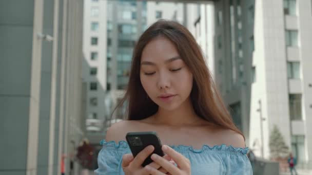 Feliz relajado joven mujer asiática sosteniendo el teléfono inteligente mirando la pantalla del teléfono celular riendo disfrutando el uso de aplicaciones móviles para llamadas de compras, charlando en las redes sociales de pie en el aire libre — Vídeo de stock