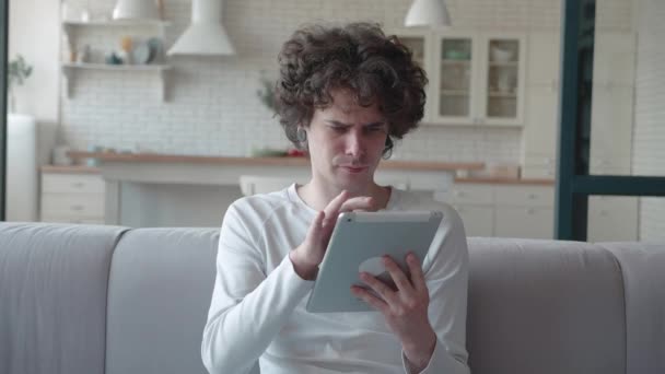 Cara surpreso lê as notícias no ipad amplia o que ele vê sentado no sofá na sala de estar — Vídeo de Stock