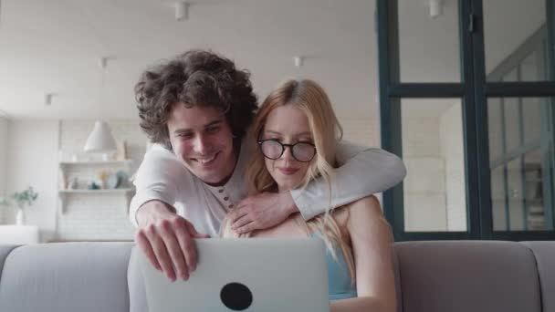 Atraktivní šťastně se usmívající blond přítelkyně pracuje na notebooku a mluví se svým milujícím radostným kudrnatým chlapem, který ji objímal kolem krku. Pečující chlap zavře notebook, aby si dívka odpočinula — Stock video
