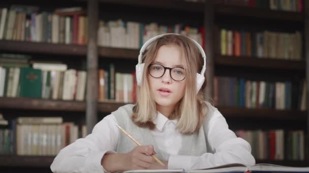 Koulutyttö oppilas yllään kuulokkeet kirjoittaa muistiinpanoja kannettavan etäopetuksen verkossa virtuaalinen oppitunti luokan opettajan kanssa kannettavan tietokoneen videoneuvottelun puhelun opiskelu kotona kirjastossa — kuvapankkivideo