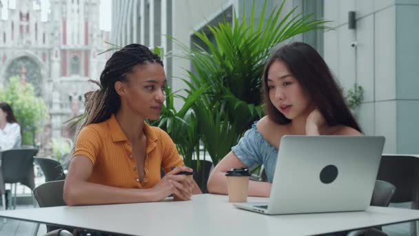 Amigas negras e asiáticas bebendo café juntas, conversando olhando no laptop e sorrindo. Jovem se senta em um café de verão e fofoca ao ar livre — Vídeo de Stock