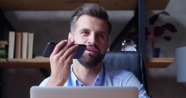 Улыбающийся бородатый молодой человек записывает голосовое сообщение на громкую связь. Бизнесмен, использующий виртуальные помощники, настраивает напоминание на смартфон в домашнем офисе. Технология цифровой и мобильной помощи — стоковое видео