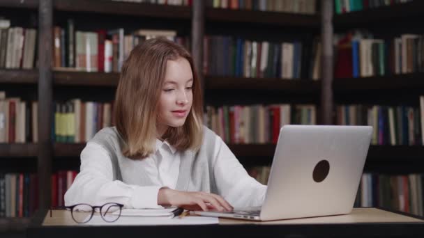 Nastolatka dziewczyna siedzi w bibliotece odpowiadając nauczyciel podczas mając lekcję online, odległość uczenia — Wideo stockowe