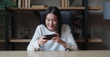 Genç Asyalı kadın akıllı telefon kullanıyor. Mobil iletişim. Masada oturan akıllı telefondan oyunu kazanan komik bir kadın.