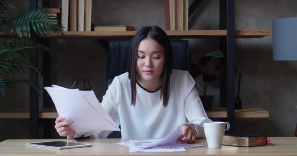 Femme asiatique assis à l'intérieur sur le lieu de travail tout en regardant à travers les documents papier dans l'appartement loft. La routine de travail — Video