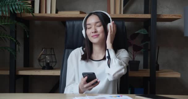 Young asian woman sitting at the table holding smartphone listening to music with headphones in modern office loft style — Stock Video