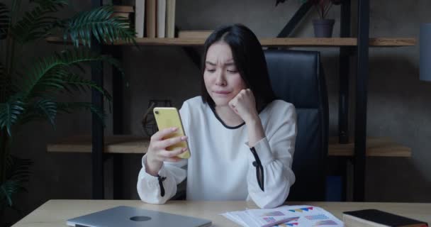 Усміхнена розслаблена азіатська жінка тисячоліття тримає смартфон, дивлячись відеоісторії соціальних мереж, сидячи на стільці за столом. Щаслива молода змішана гоночна леді, використовуючи мобільні додатки лаунж з гаджетом в офісі — стокове відео