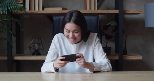 Емоційна молода азіатка грає в ігри на своєму смартфоні, сидячи на столі в квартирах — стокове відео