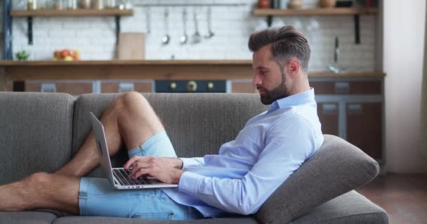 Relaxált komoly szabadúszó segítségével laptop eszköz támaszkodik kanapén otthon iroda, koncentrált vállalkozó dolgozik távolról gépelés notebook a lakásban. Fogalom vége a munkanap, támaszkodjon a kanapén — Stock videók
