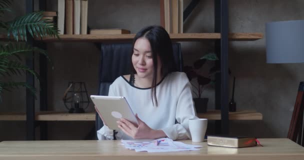 Ділова жінка сидить за столом в офісі, використовуючи планшет. Молодий підприємець посміхається, серфінг в Інтернеті або працює — стокове відео