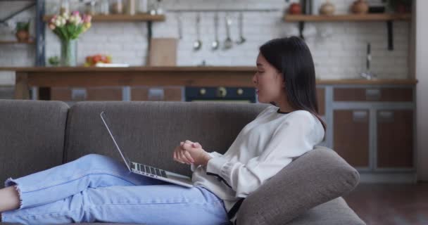 Mujer asiática que utiliza el ordenador portátil para el chat de vídeo en el sofá fondo de cocina. — Vídeos de Stock