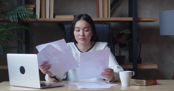 アジアの女性オフィスマネージャーは、ビジネス文書の膨大な山をソートし、ラップトップ画面を見ています。白いシャツを着た女性がテーブルの上に腰を下ろしている。 — ストック動画