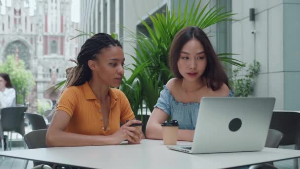 Rase misto e asiático amigos do sexo feminino beber café juntos, falando olhando no laptop e sorrindo. Jovem se senta em um café de verão e fofoca ao ar livre — Vídeo de Stock