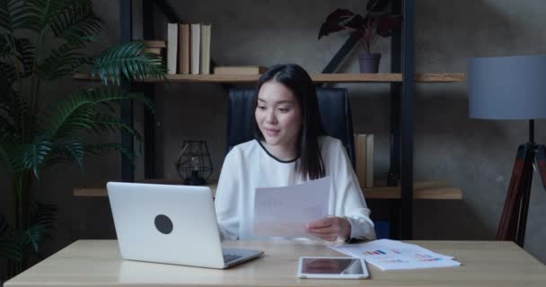 Zelfverzekerde Aziatische vrouwelijke web coach, online leraar of customer support service operator spreken tot webcam kijken naar laptop weergegeven diagram op papier. Zakelijke vrouw maken conferentie video call concept — Stockvideo