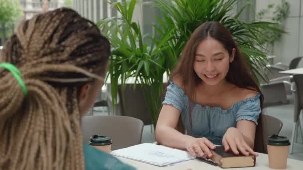 Enfoque mujer joven asiática con libro sus notas. Estudiantes universitarios amigos aprendiendo y enseñando juntos en un restaurante. Sobre el hombro disparado — Vídeos de Stock
