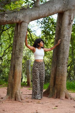 Bir kadın iki ağacın önünde poz veriyor, beyaz bir bluz ve leopar desenli pantolon giyiyor.