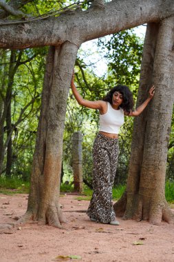 Bir kadın iki ağacın önünde poz veriyor. O, beyaz bir atlet ve siyah pantolon giyiyor. Ağaçlar uzun ve bir sürü yaprakları var. Kadın ağaçlardan birine yaslanıyor.