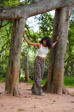 Bir kadın iki ağacın önünde poz veriyor, biri diğerine yaslanıyor. O, beyaz bir bluz ve siyah pantolon giyiyor. Bu görüntünün doğal ve huzurlu bir havası var.