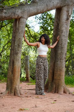 Bir kadın iki ağacın önünde poz veriyor. Beyaz bir bluz ve leopar desenli pantolon giyiyor. Resmin doğal ve huzurlu bir havası var. Ağaçların ortasında duran kadın.
