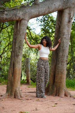 Ağacın önünde beyaz bir atlet ve leopar desenli pantolon giymiş bir kadın duruyor.