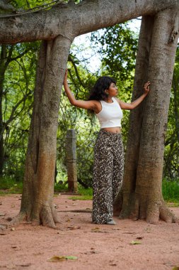 Bir kadın iki ağacın önünde poz veriyor. O, beyaz bir bluz ve siyah pantolon giyiyor. Ağaçlar uzun ve bir sürü yaprakları var. Kadın ağaçların ortasında duruyor.