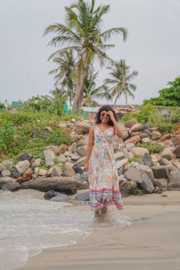 Plajda beyaz bir elbise giymiş bir kadın yürüyor. Plaj kayalıktır ve arka planda palmiye ağaçları vardır. Kadın gülümsüyor ve kumsalda eğleniyor.