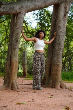 Bir kadın iki ağacın önünde poz veriyor, leopar desenli pantolon ve beyaz bir bluz giyiyor.